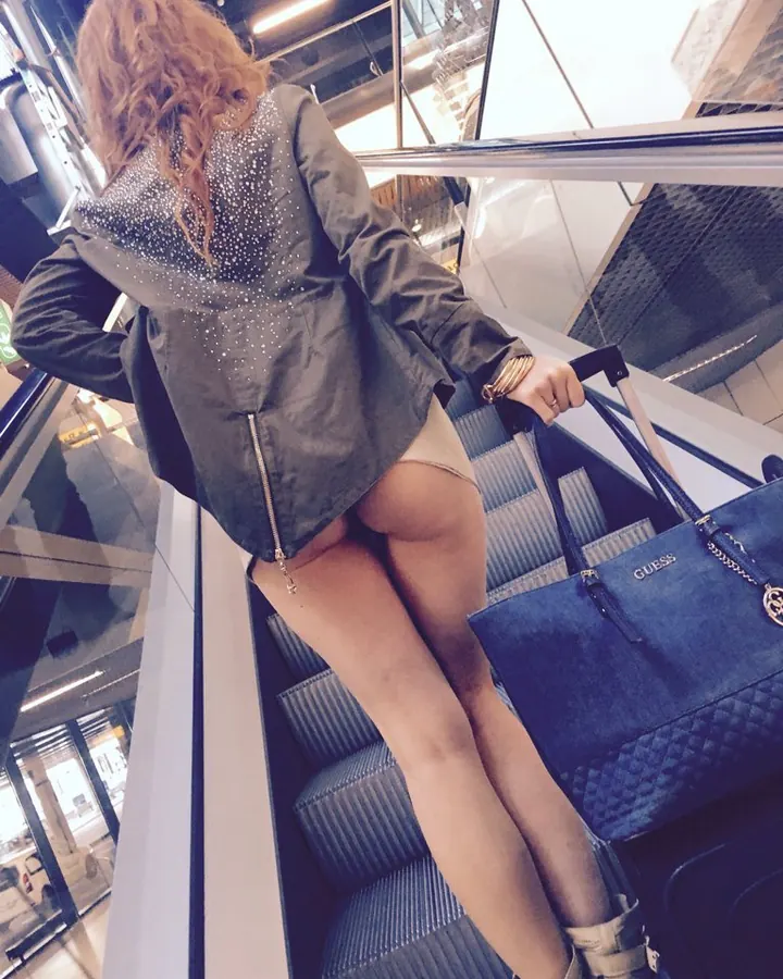 Femme aux cheveux châtains en veste noire pailletée, vue de dos sur un escalator, tenant une valise bleue