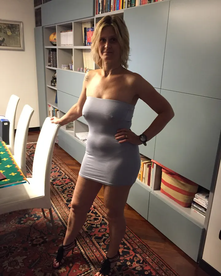 Jeune femme aux cheveux blonds en robe grise moulante, debout dans un salon avec un tapis oriental et une bibliothèque