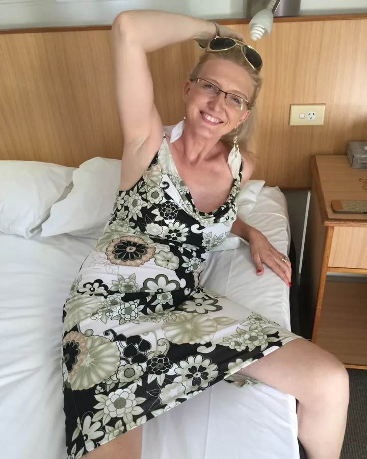 Femme blonde aux cheveux mi-longs, en haut noir, posant dans une chambre.