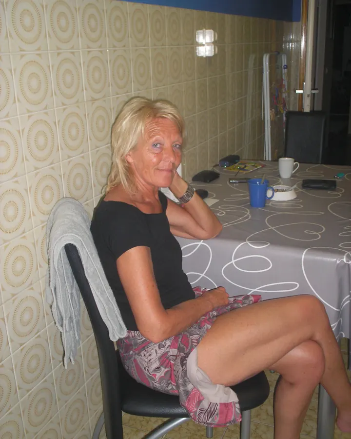 Femme blonde aux cheveux courts, en t-shirt noir, assise à table, regardant vers la caméra.