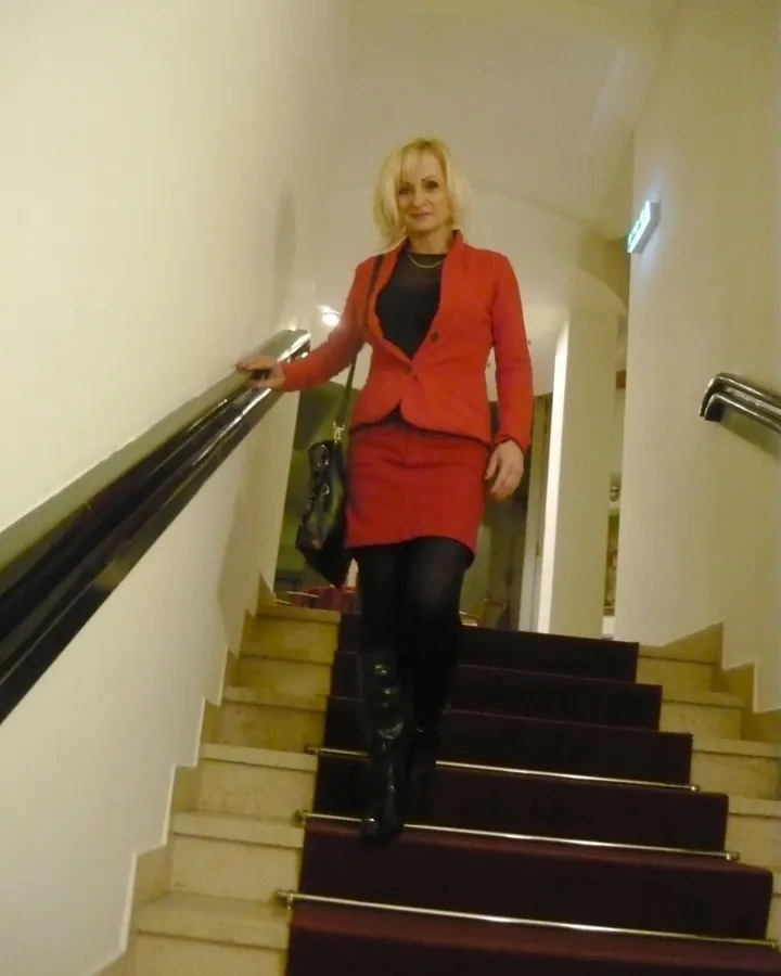 Femme blonde en tailleur rouge descendant un escalier intérieur, tenant une poignée métallique.