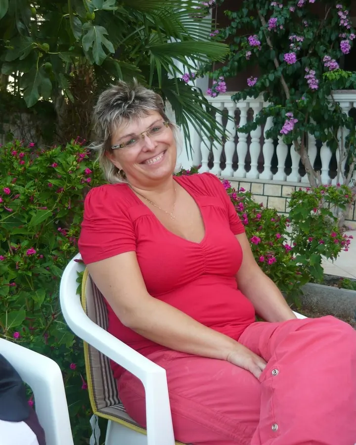 Femme en robe rouge, assise sur une chaise blanche, souriante, entourée de fleurs roses et violettes.