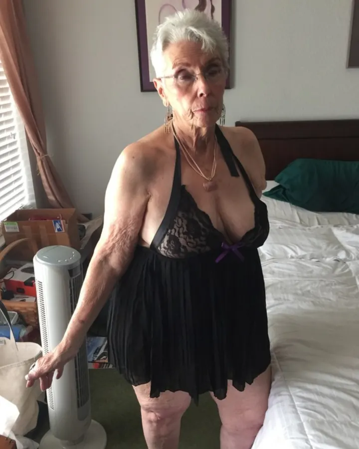 Femme aux cheveux blancs, debout dans une chambre, portant une robe noire à dentelle et un collier.
