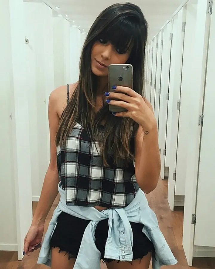 Femme aux cheveux bruns-noirs, prenant un selfie dans un couloir, portant un haut à carreaux et un short.